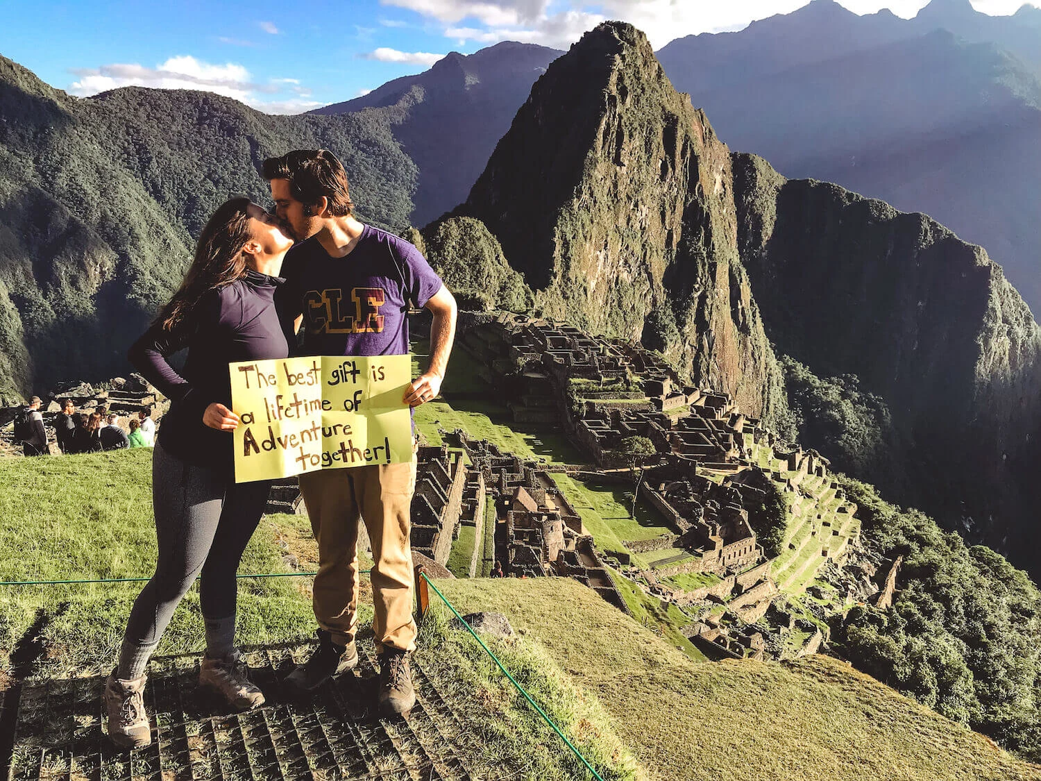 Kat and Chris holding a sign saying -The best gift is a lifetime of adventure together,- while kissing- overlooking Machu Picchu - Peru itinerary