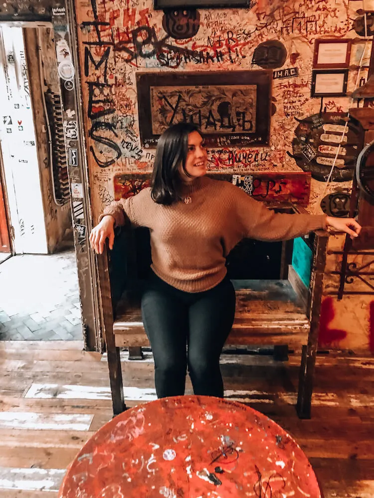 Kat wearing black trousers and a brown sweater sitting in a chair next to a graffiti wall at Szimpla Kert in Budapest, Hungary