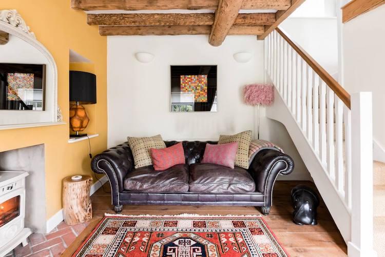 Cotswolds Airbnb | Living Room of a cottage with wood beams, yellow fireplace accent wall, and cozy leather couch