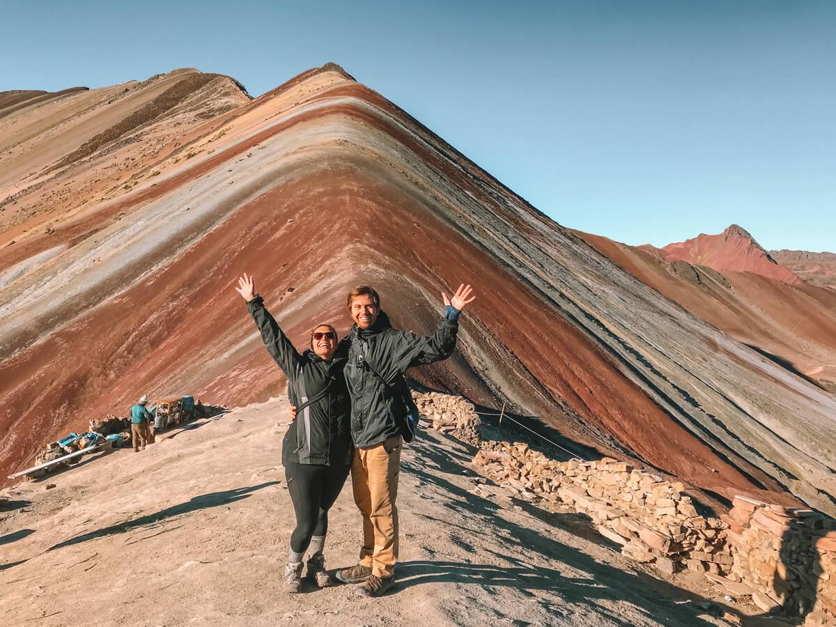 Episode 100 Top Travel Memories and Rapid Fire Travel Questions - Kat and Chris smiling and waving at the end of the Rainbow Mountain Trek