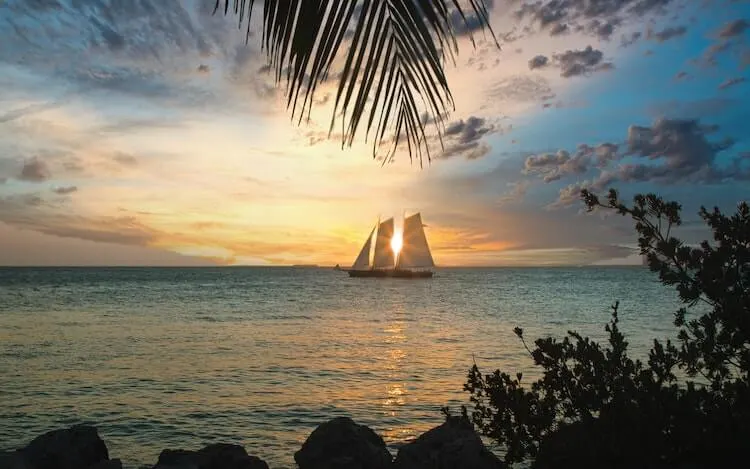 Key West, Florida at sunset with a sailboat in front of the sun and sun peaking through - US Honeymoon Destinations