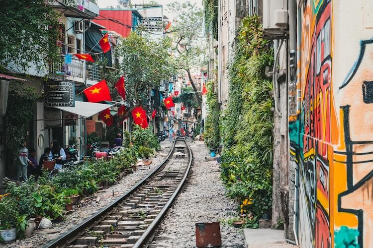 Railway street in Vietnam with plants growing beside it and home and stores with Vietnamese flags on the left