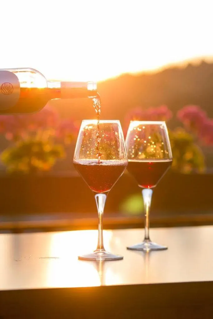Red wine being poured into a glass as it sits beside another glass with the sunset over a vineyard in the background