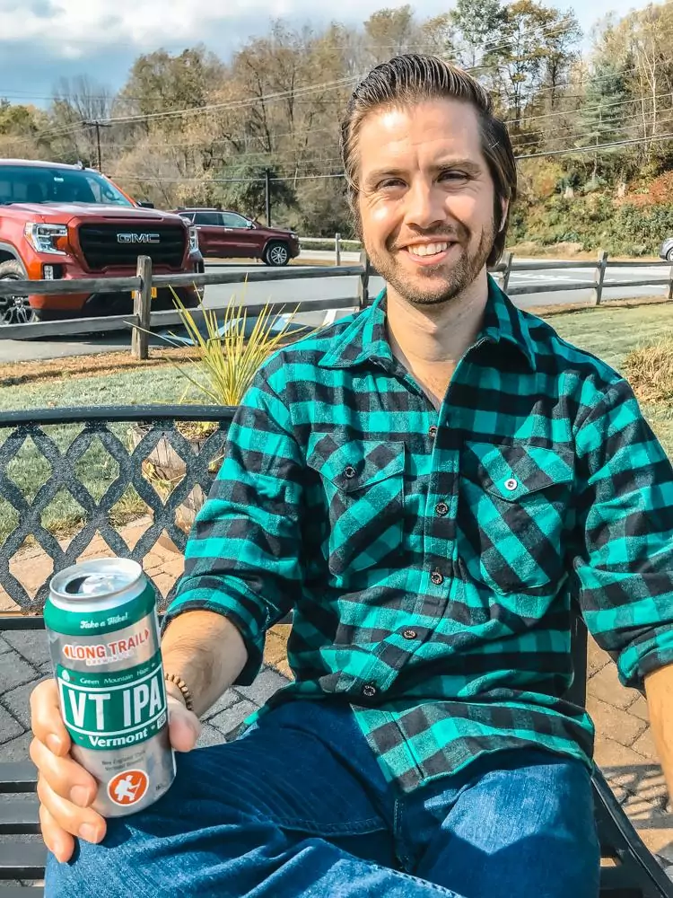 Chris in a green and black flannel and jeans sitting with his leg crossed on his knee and holding a Vermont IPA