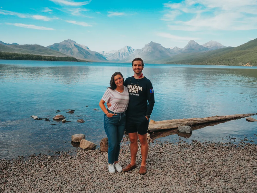 Kat and Chris with an arm around each other and smiling at the camera with Lake McDonald in the background with mountains further back