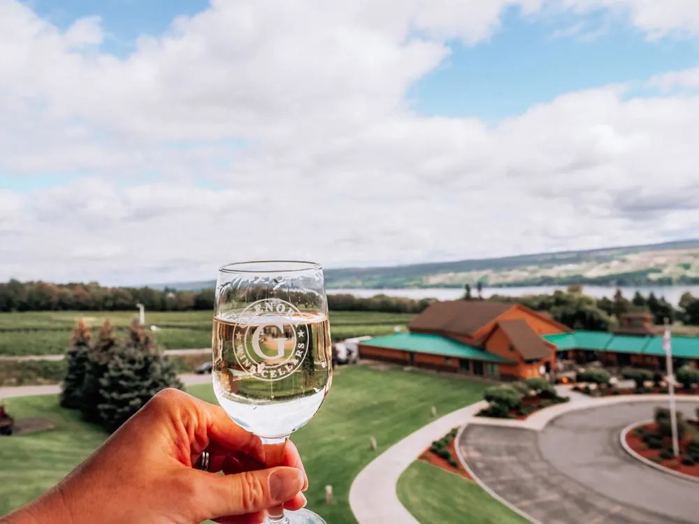 Glenora Cellars | Finger Lakes Romantic Getaway | White wine in a glass being held by a hand with a lodge and lake in the background