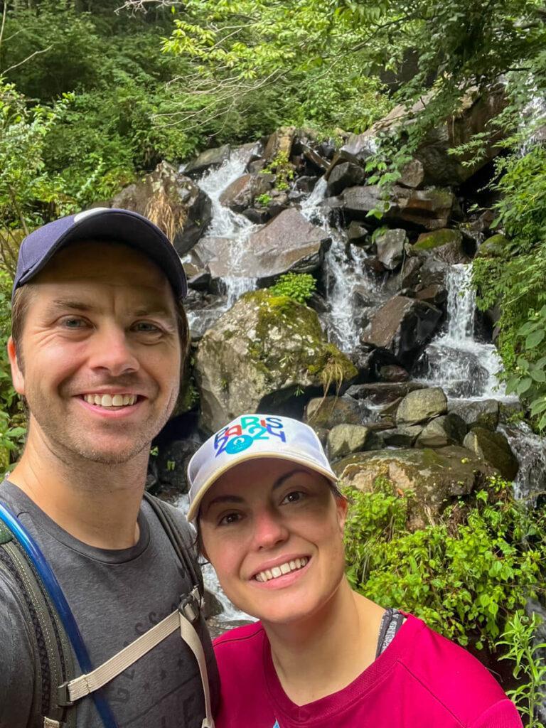 Kat and Chris smiling at the camera with a waterfall in the background while hiking in Hakone
