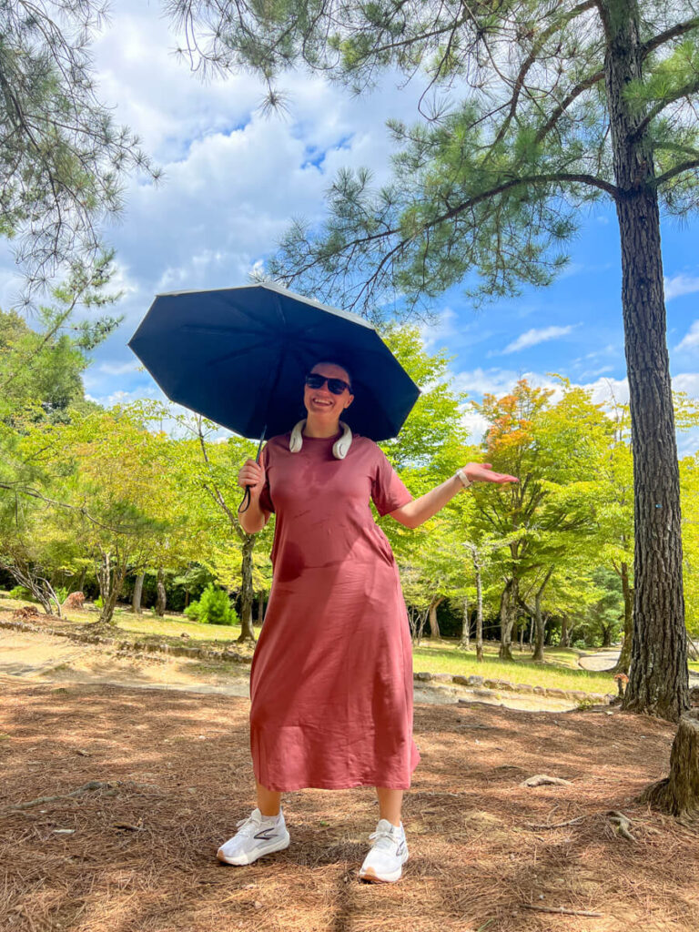 Kat smiling while standing under her UV umbrella with her neck fan on and sweat on her pink dress