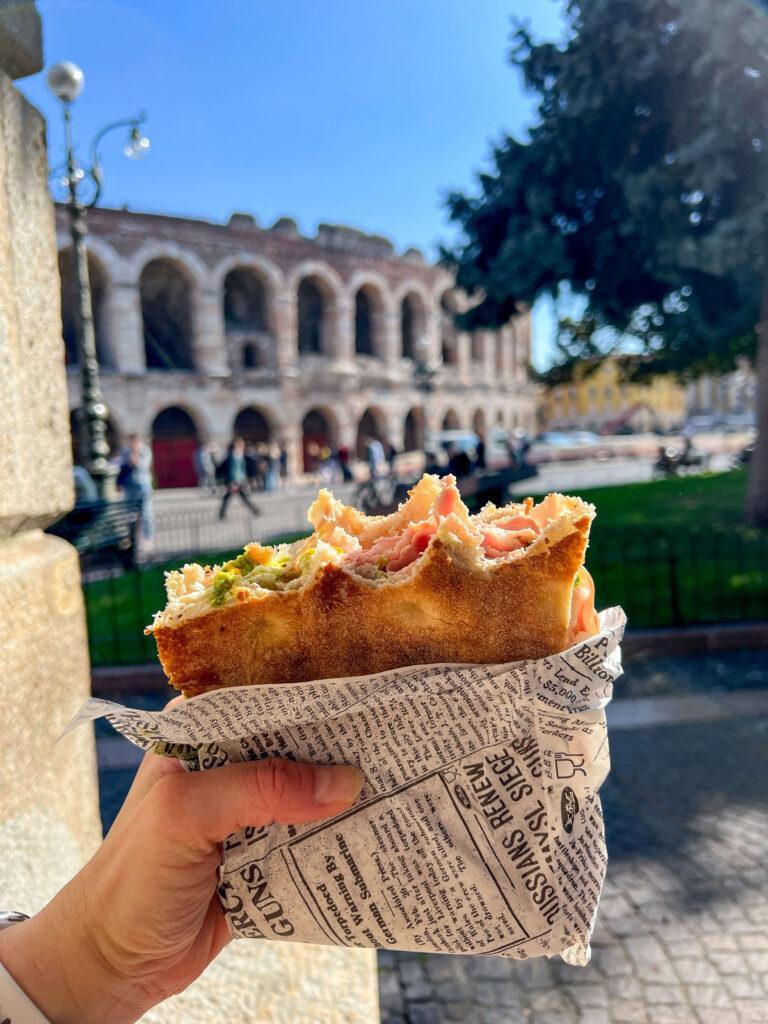 Panini from Brusco Panini with a few bites taken from it being held up with the Arena di Verona in the background